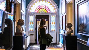 A visitor stands in a hallway with numerous paintings and busts of suffragists.