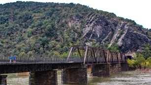 Two hikers on a bridge over a river leading to a tunnel in a mountain