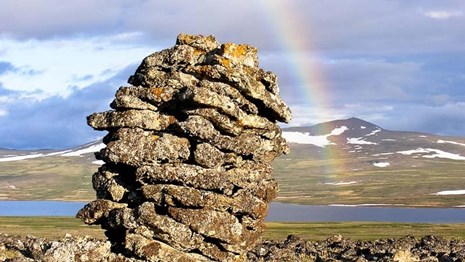 Tall rock cairn with rainbow over a lake and mountains in the distance