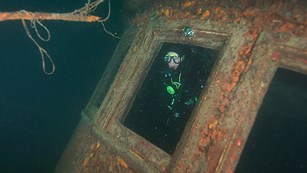 man in scuba diving wet suit underwater looking through a window of a shipwreck