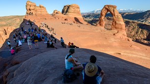A crowd of visitors gathers near Delicate Arch with views of mountains.