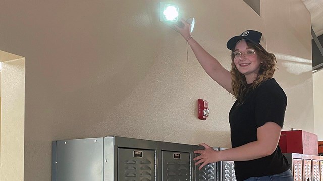 A woman on a ladder reaches up to a safety light above lockers.