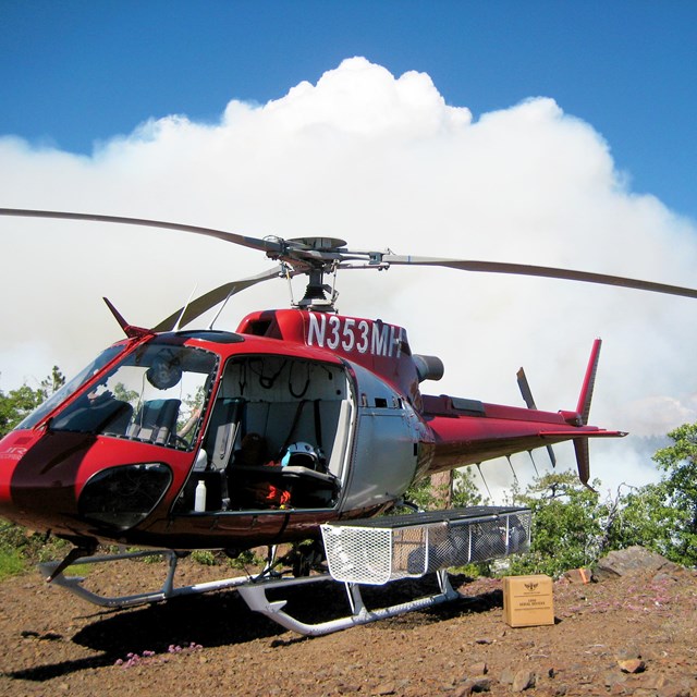 A helicopter stages during a prescribed burn a smoke column is in the background.