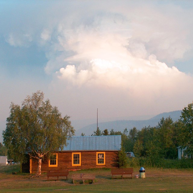 A wildfire's smoke column is off in the distance and a wooden cabin in is the foreground.