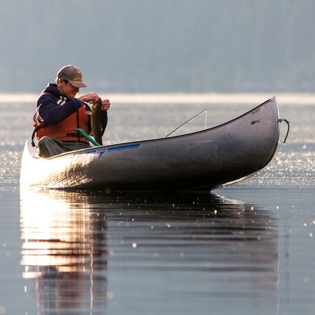 man in canoe on water holding a newly caught fish. 