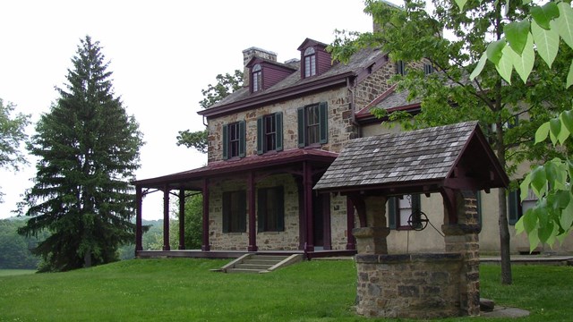 View of green lawn and a 19th century stone house with maroon trim and a covered well.