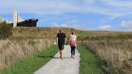 Two visitors walking a trail uphill toward the visitor center and an elevated overlook.