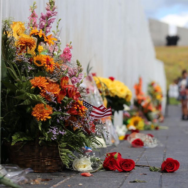 Bright flowers spilling from a basket with others along a white memorial wall, visitors walk beside.