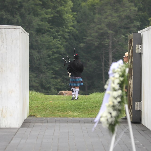 Wreath by the Wall of Names, a babpiper walks out to the boulder through open Ceremonial Gate.