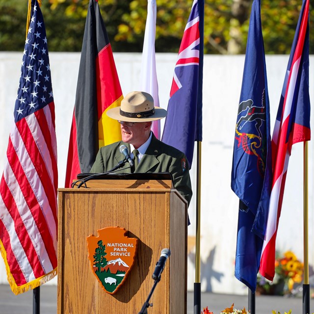 Park Ranger speaking in front of a podium with flags behind.