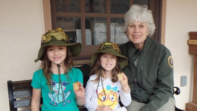 An adult and two kids sit on a bench, holding activity books and a junior ranger badges.