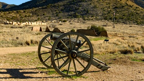 A howitzer is in the foreground; ruins of walls and mountains are in the background.