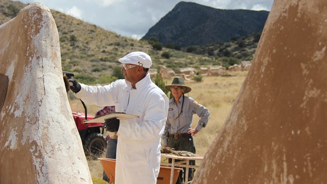 A man is applying plaster to the ruins of a wall.  Other workers and mountains are in the background