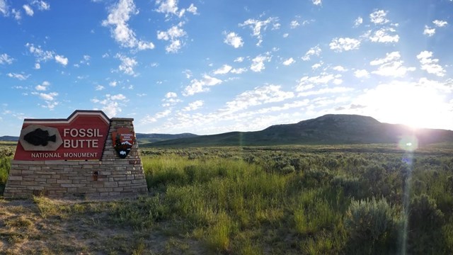 Sign that says Fossil Butte National Monument with fossil fish and arrowhead. Sun rising over butte.