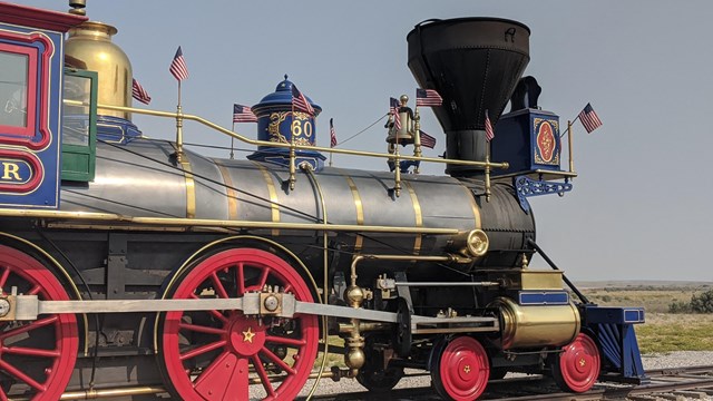 The front of a train engine facing right painted red, blue, gold, and black
