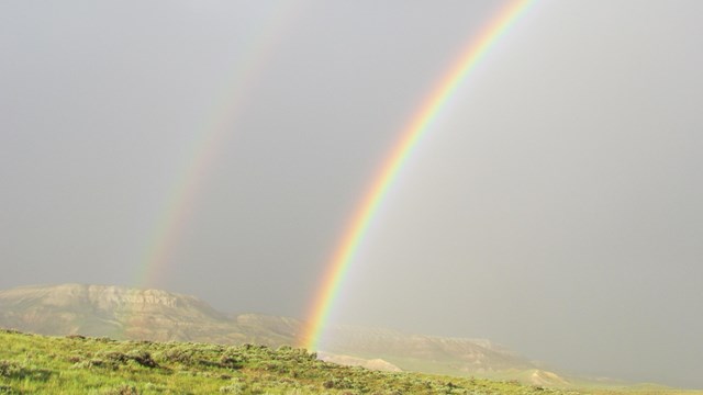 A double rainbow arcs through a grey sky with a bright green hill below.