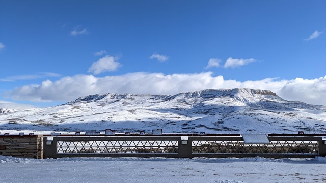 A snow-covered porch with a snow-covered butte behind. The sky is bright blue with some clouds.