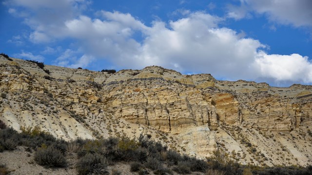 A cliff with yellow, brown, and gray horizontal layers with a blue sky and clouds above.