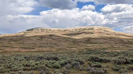 Fossil Butte with the top in sunshine and the bottom in shade. A cloud-spotted blue sky is above.