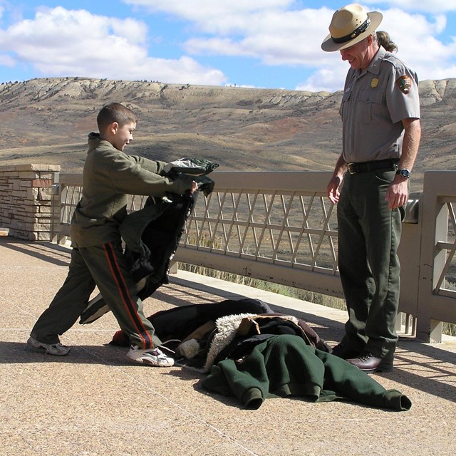 A child places a coat on top of a pile of coats while a ranger watches.