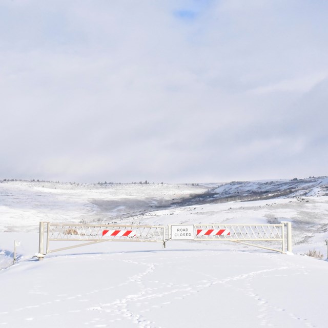 A gate with a sign saying road closed blocks Fossil Butte's scenic drive which is covered in snow.