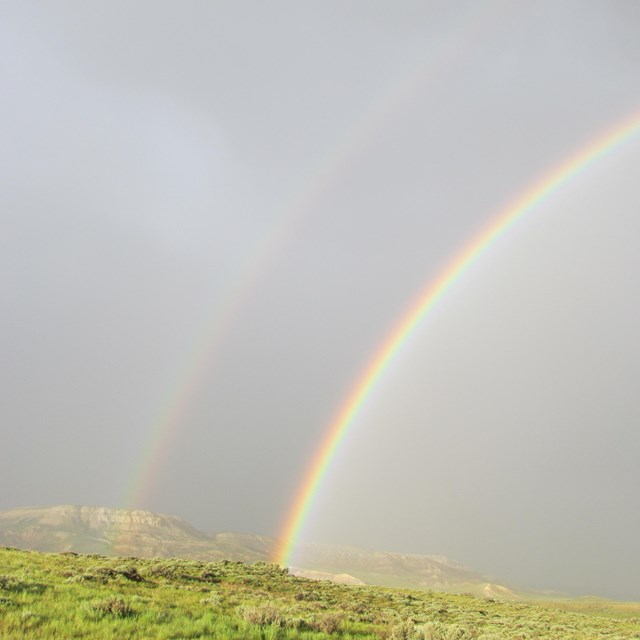 A double rainbow arcs through a grey sky with a bright green hill below.