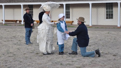 A soldier in blue wool outfit on one knee proposing to his future bride and her daughter.