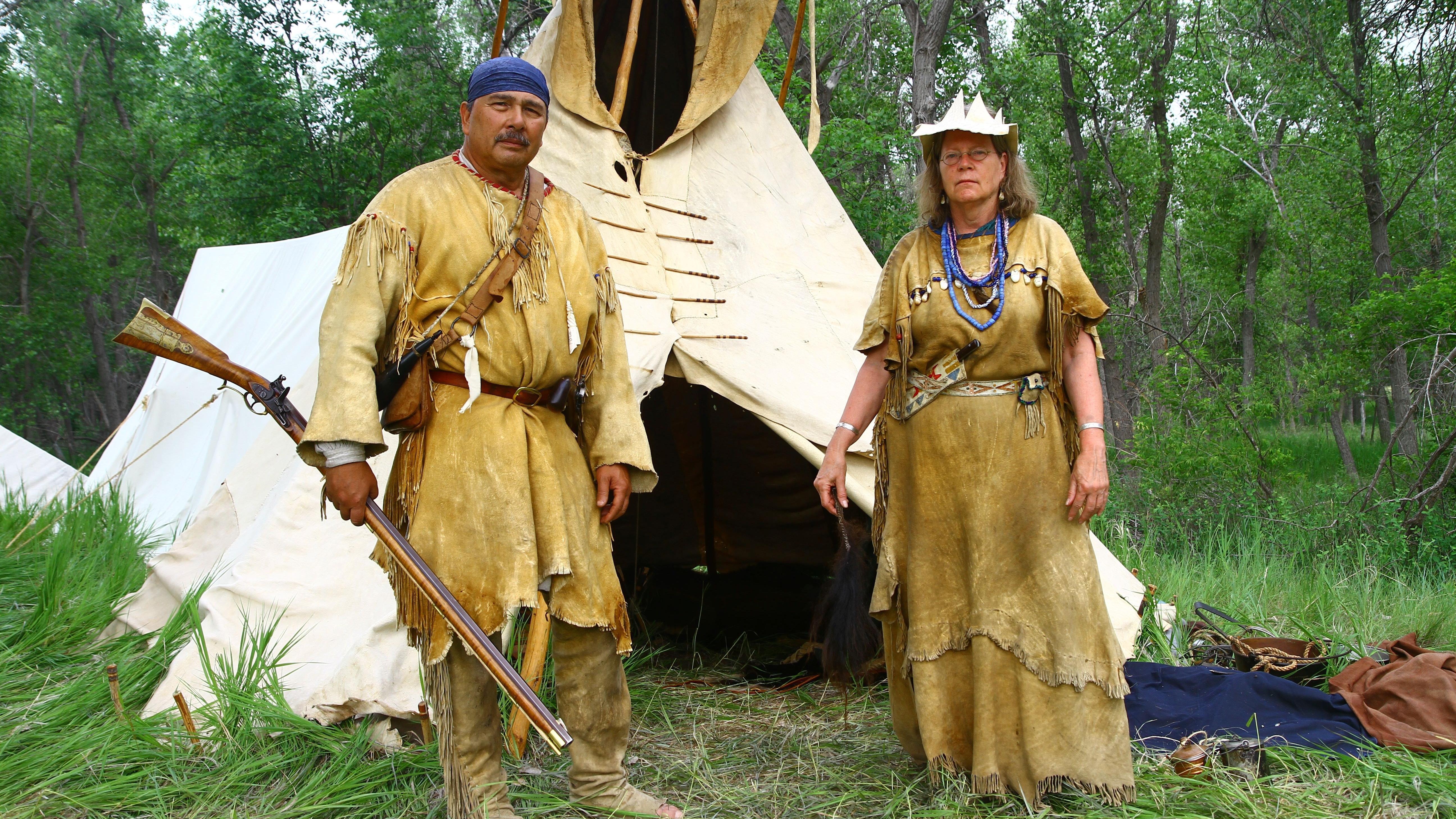 Two individuals in traditional attire stand beside a teepee, showcasing cultural heritage
