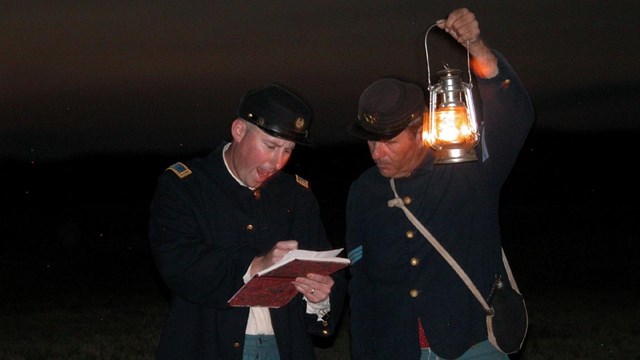 Image of two men dress as uniformed soldiers looking at piece of paper by lantern light.