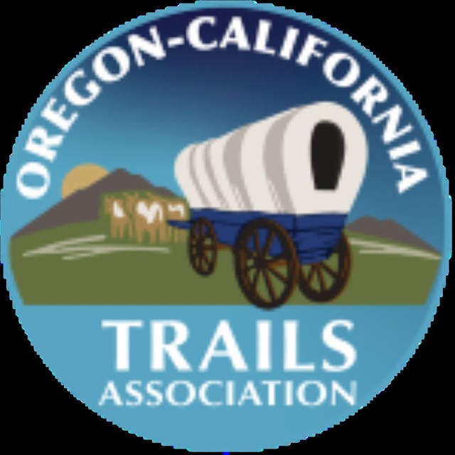 A covered wagon sits in front of a cow and a mountain. 