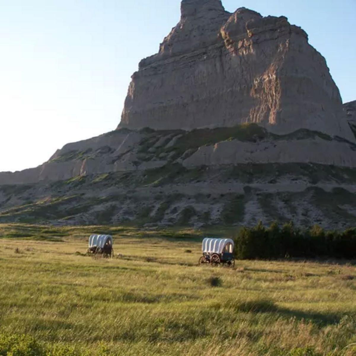 A large, rugged rock formation rises prominently against a clear blue sky, with wagons. 