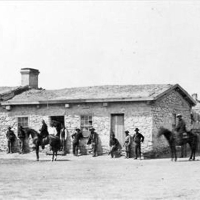 Black and White image from the 1800s. Several people standing outside an adobe style small building