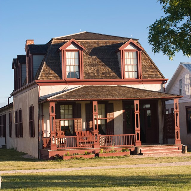 Large house made out of brown adobe structure, with red wood trim, and a gray roof. 