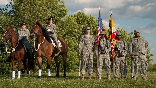Three members of the military standing with flags, and two sitting on horses.