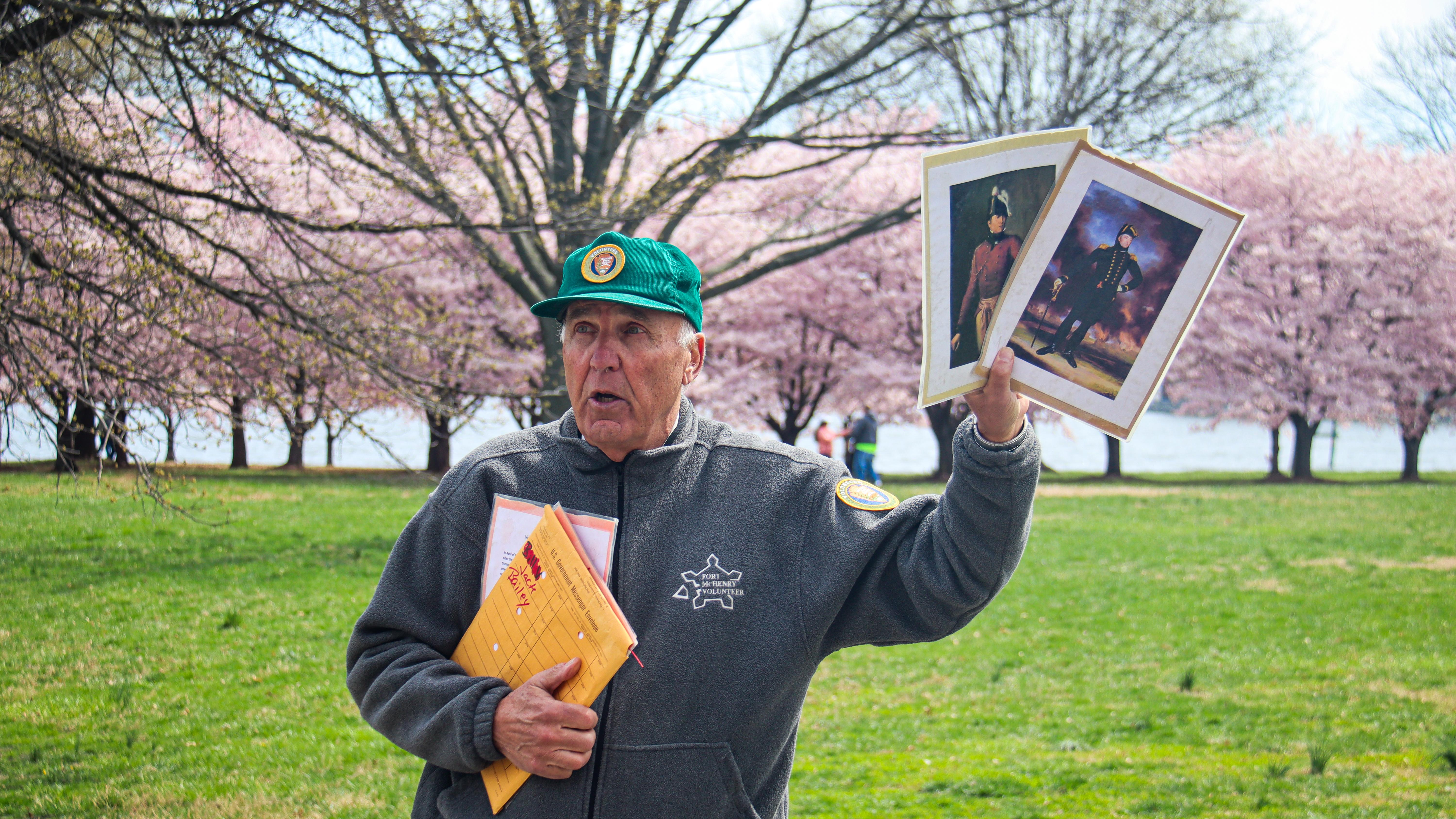 NPS volunteer in a green hat and grey jacket holding up two images as he speaks in a grassy field.