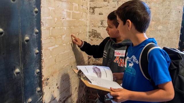 Students stand in front of a brick wall inside Fort Point while holding journals