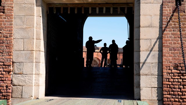 A man in period clothing pointing something out to a group of 3 people, seen through a large doorway