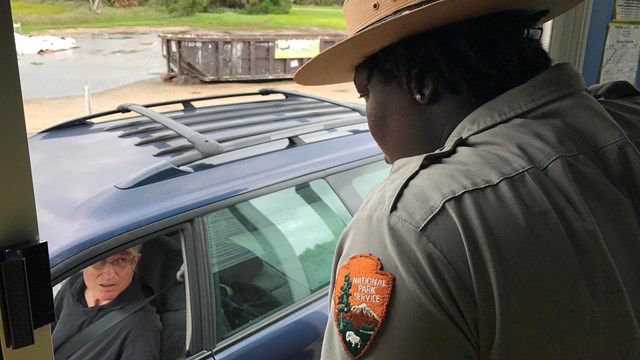 A park ranger standing in a window talking to a man in a car. 