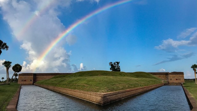 A rainbow above a brick fort with a moat in front. 