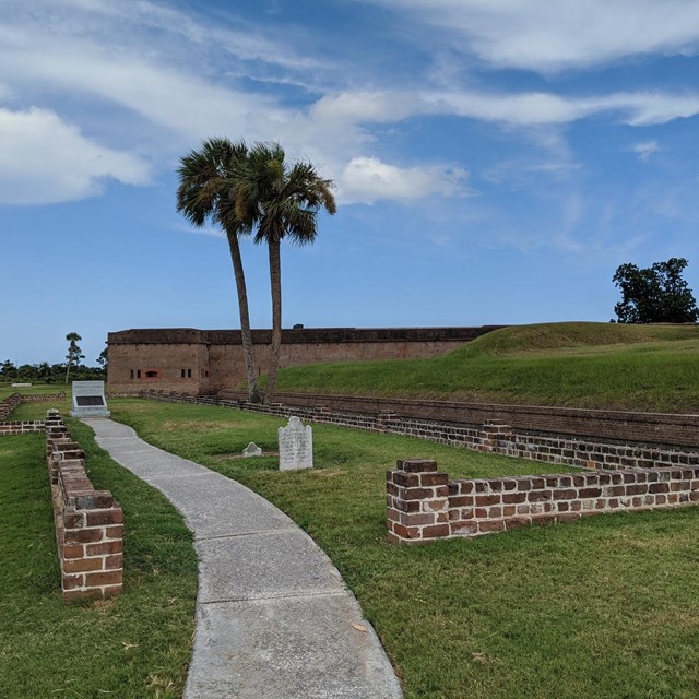 concrete path through cemetery with three tombstones, brick fort in background.