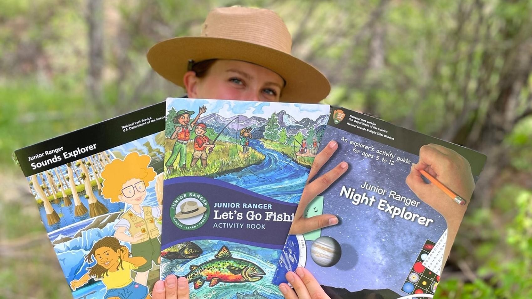 A ranger holding three different junior ranger booklets obscuring the lower half of their face.