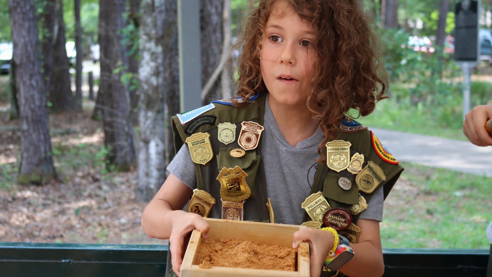A child in a junior ranger vest carrying some sand in a wooden container