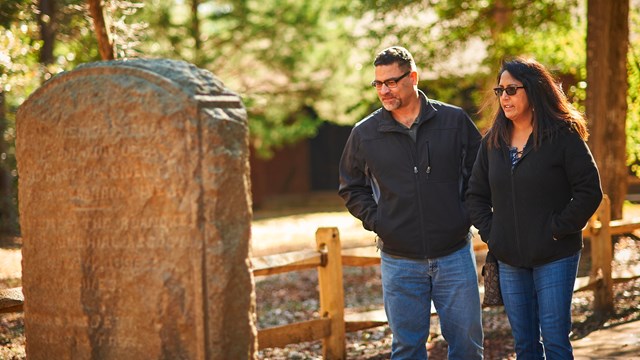 Two visitors look at a vertical stone monument with green forest areas behind them