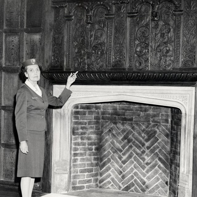 Black and white photo of a woman showing off carvings over a fireplace
