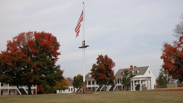 The grassy parade ground with trees, american flag flying on the flag pole, and officer's quarters.