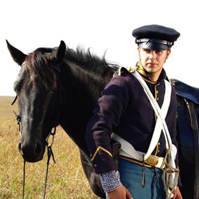 An interpreter dressed as a 19th century U.S. Dragoon standing in front of horse. 