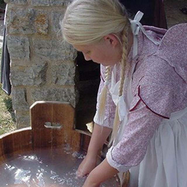A girl in 19th Century dress launders clothes in a wooden bucket