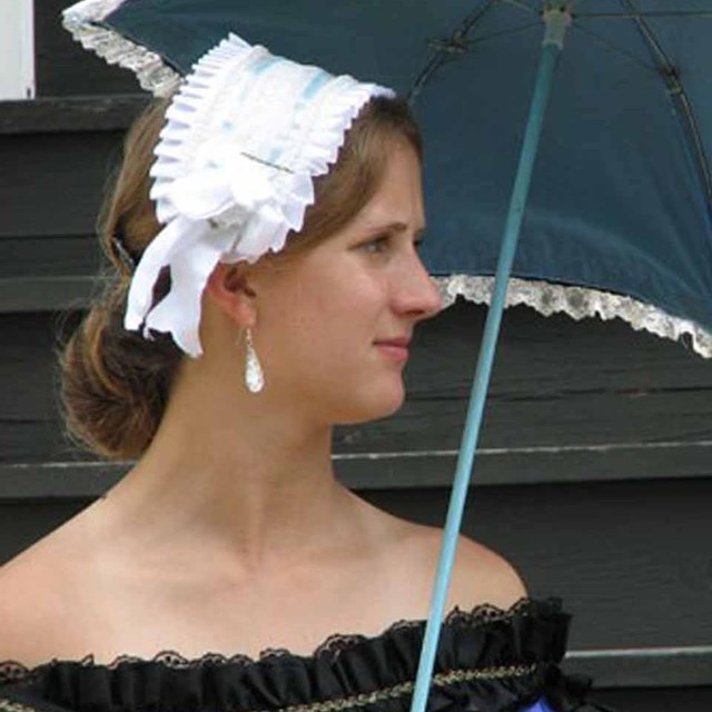 A woman in period dress holding a parasol stands in front of stairs to the officer's quarters