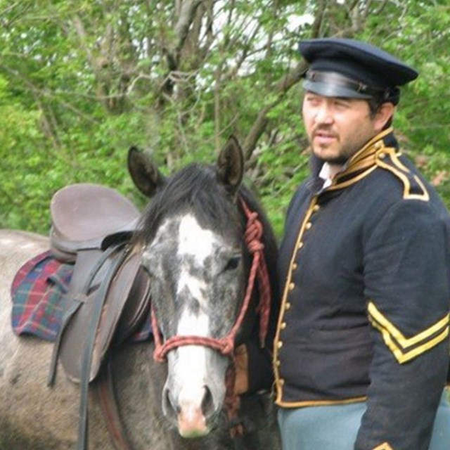 A man in US dragoon dress stands with a tacked horse