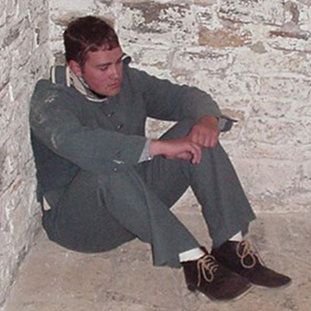 A man in soldier's dress slumps in the corner of a guardhouse cell